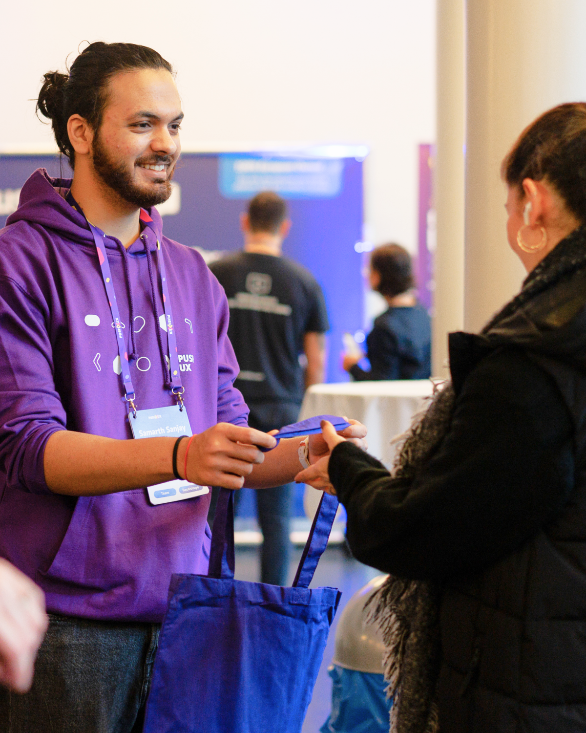 Smiling PUSH UX volunteer in a purple hoodie handing a tote bag to an attendee at the venue.